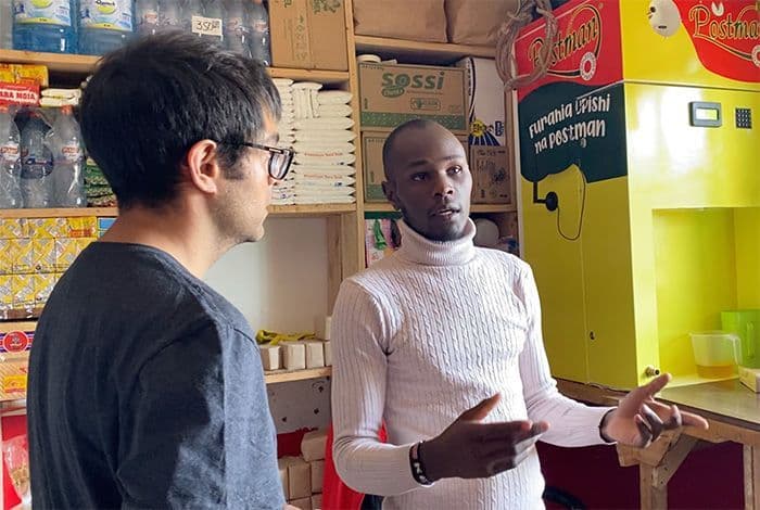 Two men discuss in a shop, one gesturing while speaking to the other.