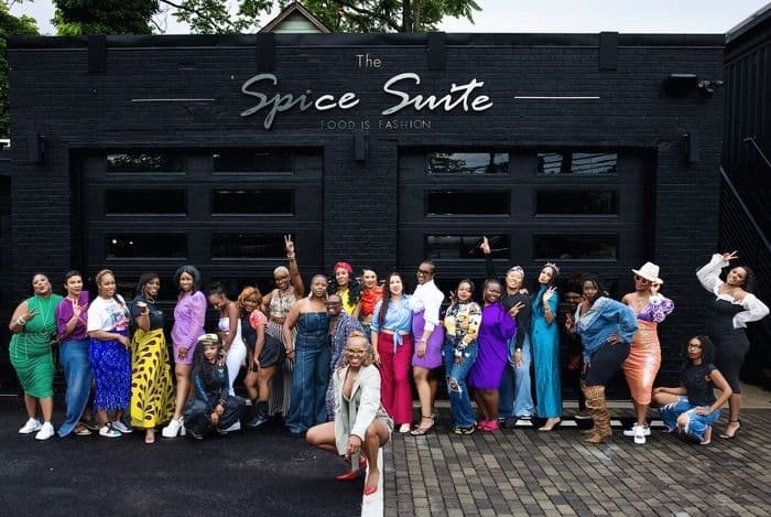 A large group of diverse women in colorful outfits pose in front of The Spice Suite building.